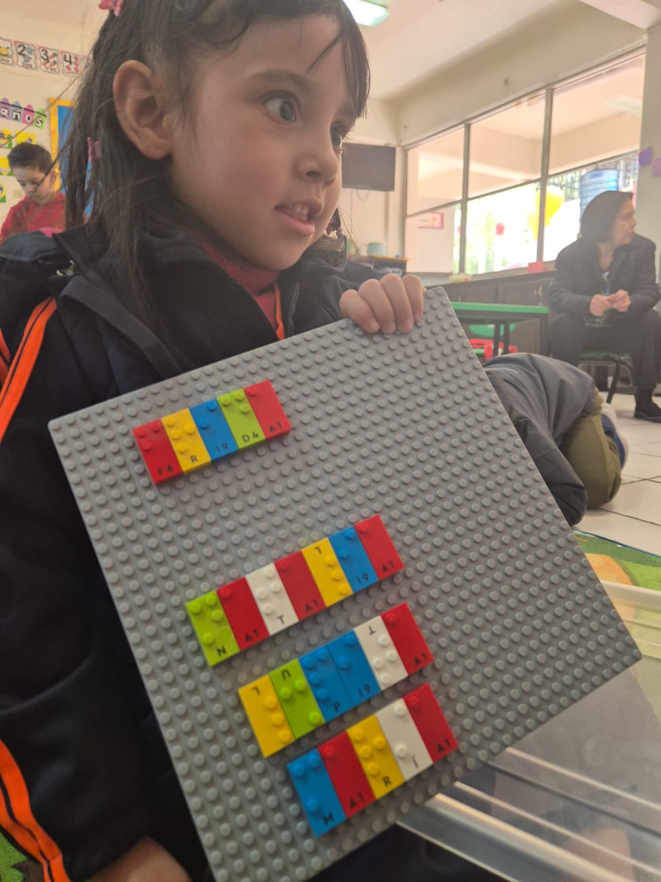 Natalia María sostiene una placa base LEGO gris con LEGO Braille Bricks de colores dispuestos en filas, cada uno de ellos con letras, números y colores. Está en el interior de un aula con otras personas al fondo.