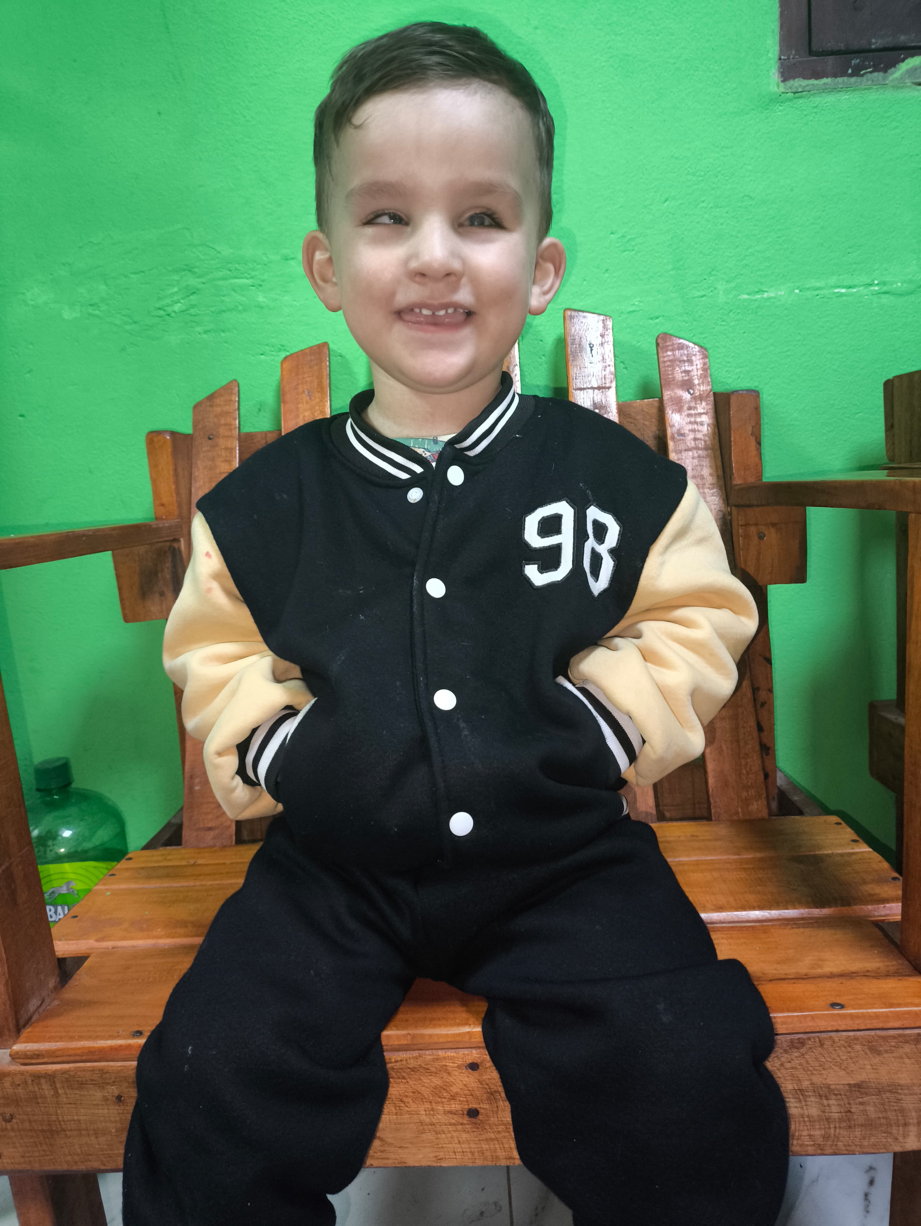 Un joven sonriente, vestido con una chaqueta negra y beige con la inscripción "98" en la parte delantera, está sentado en un banco de madera contra una pared verde brillante.