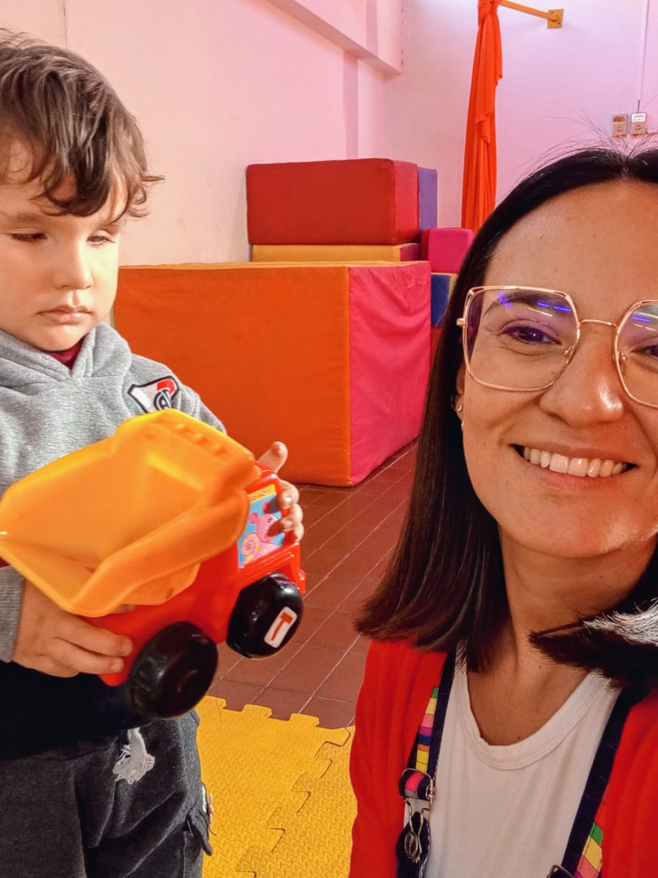 Una maestra con gafas y el pelo negro sonríe a la cámara mientras Naún sujeta un camión volquete rojo de juguete. En el fondo se ven bloques de espuma de colores y una colchoneta amarilla.