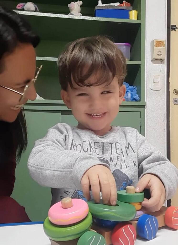 Naún sonríe mientras juega con coloridos juguetes de madera en una mesa, mientras una maestra a su lado le observa con una cálida sonrisa. Hay estanterías y una puerta al fondo.
