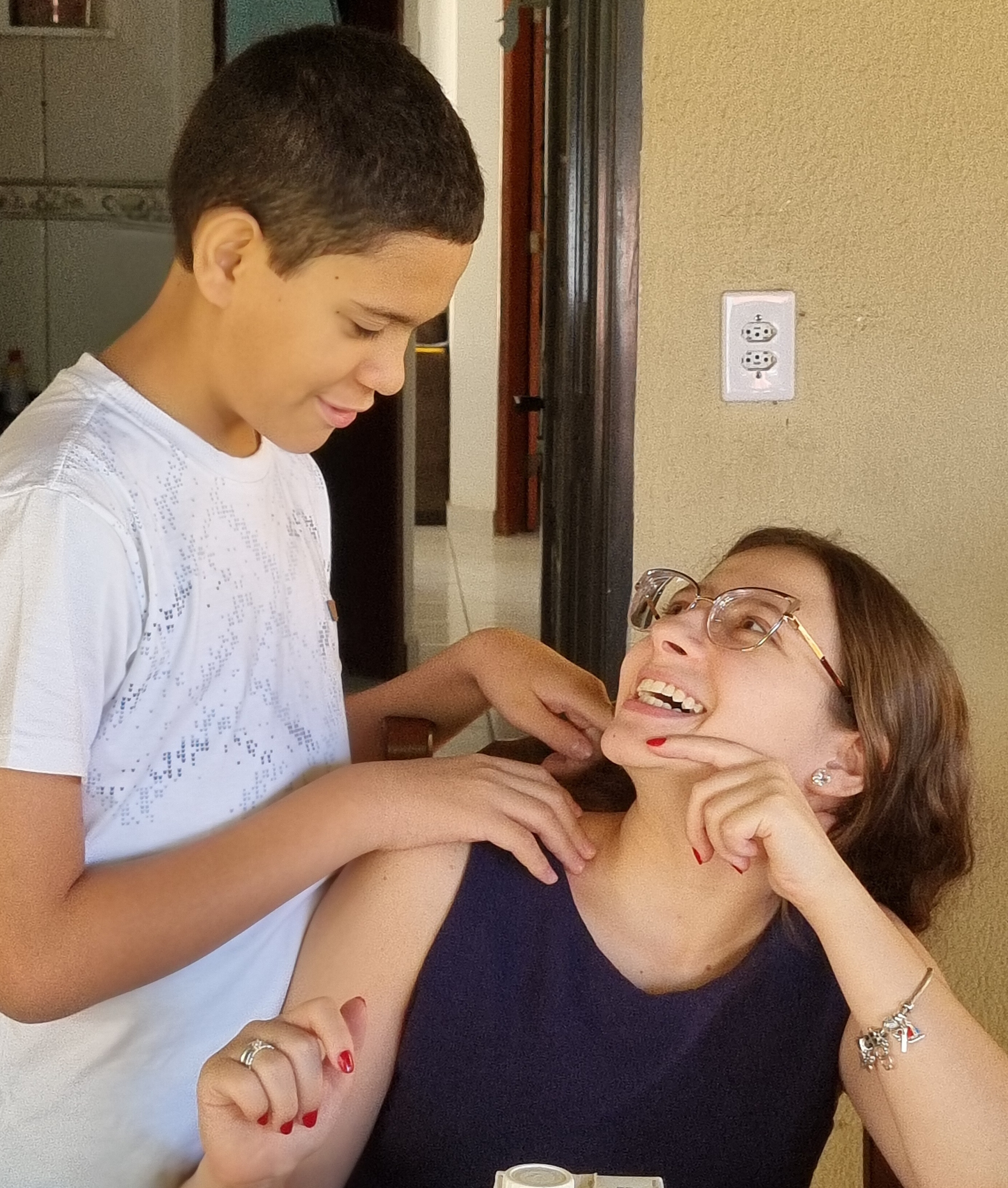 Sarah, uma jovem sorridente de óculos, está sentada à mesa e olha para Pedro, que está ao lado dela, tocando-lhe gentilmente os ombros. Ambos parecem felizes e envolvidos, partilhando um momento alegre dentro de casa.