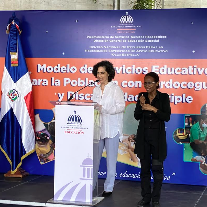 Una mujer habla en un podio mientras otra mujer está de pie a su lado haciendo señas. Detrás de ellas hay una pancarta sobre los servicios educativos para personas con sordoceguera y discapacidad múltiple en la República Dominicana. Se ve la bandera dominicana.