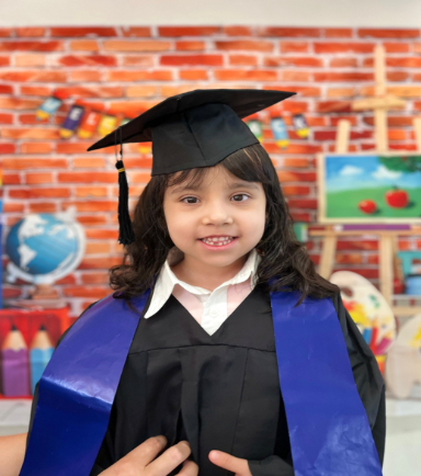 Una joven con toga y birrete de graduación sonríe delante de un colorido fondo de clase con un globo terráqueo, lápices y un caballete.