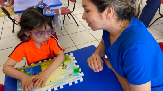 Natalia María, una joven con cabello oscuro y anteojos morados, juega con un juego de mesa táctil en una mesa azul, mientras una maestra se sienta a su lado, guiándola e interactuando con ella atentamente.