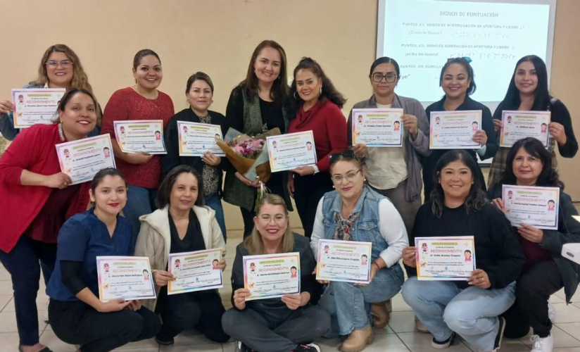  Un grupo de maestras sonrientes y con certificados en la mano posan juntas en un interior; una mujer en el centro lleva un ramo de flores. Al fondo se ve una presentación proyectada.