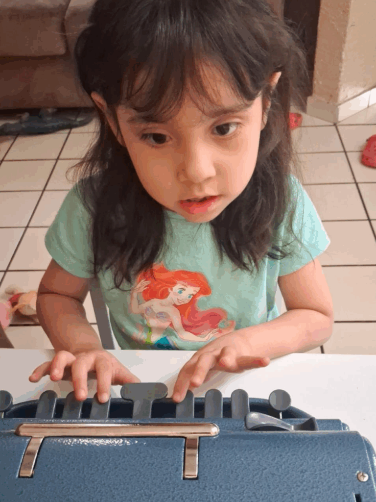 Natalia María con una camiseta de La Sirenita utiliza una máquina Perkins Brailler en una mesa, centrándose en sus manos. El fondo muestra un sofá, suelo de baldosas y juguetes esparcidos.
