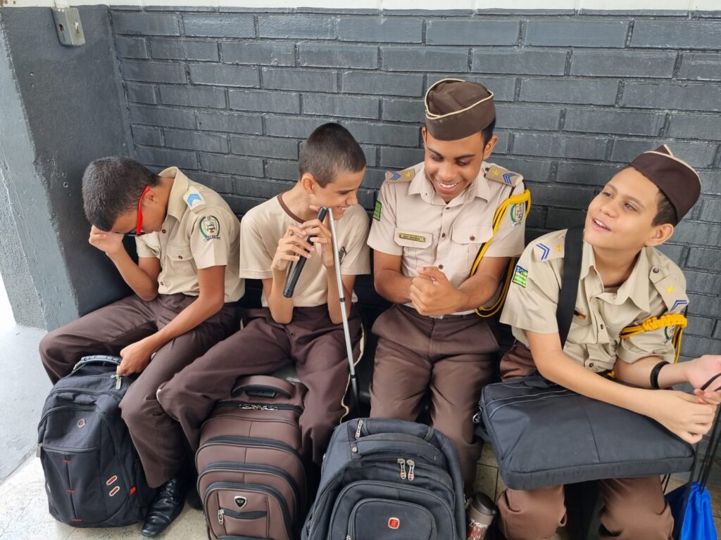 Quatro meninos com uniformes e chapéus marrons combinando sentam-se em um banco contra uma parede de tijolos cinza, sorrindo e rindo juntos, segurando suas bolsas no colo.