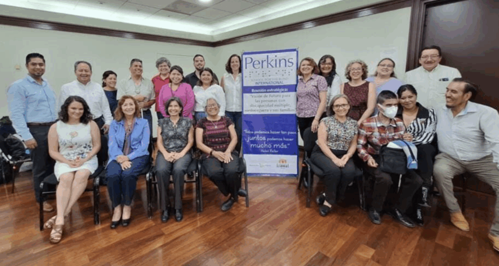 Un grupo de unas 20 personas posa y sonríe para una foto en una sala de conferencias. Detrás de ellos hay una pancarta azul con el logotipo de Perkins y texto. Algunas personas están sentadas, mientras que otras permanecen de pie al fondo.
