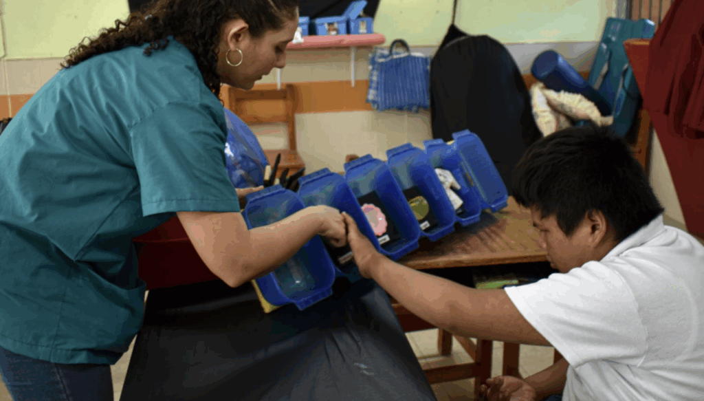 Una mujer ayuda a un niño sentado a interactuar con objetos de colores dentro de cubos azules dispuestos en fila sobre una mesa en un aula.