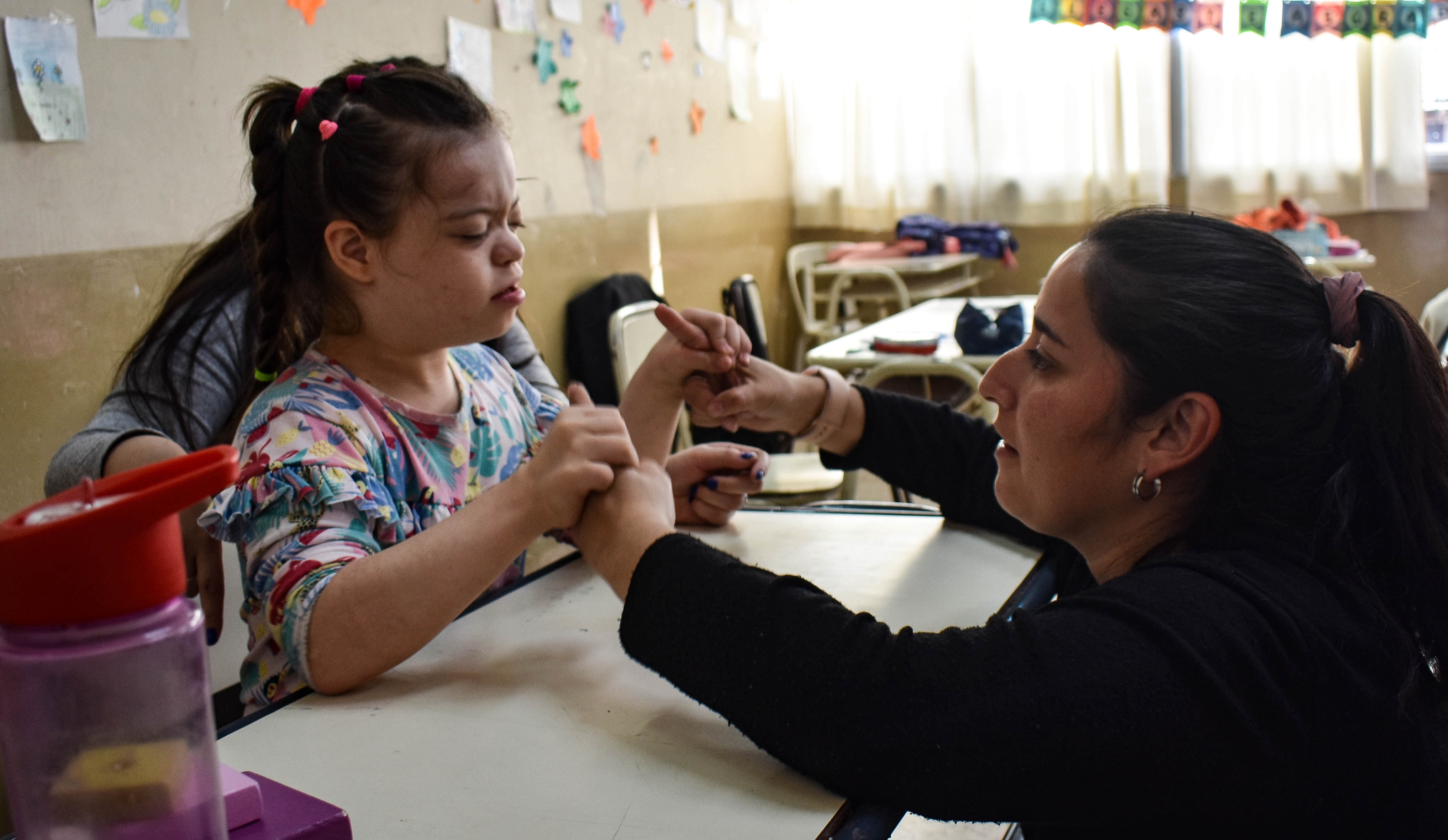 Una docente interactúa con una niña en un aula muy iluminada, sosteniéndole suavemente las manos sobre un pupitre. En las paredes del fondo se ven dibujos y adornos.