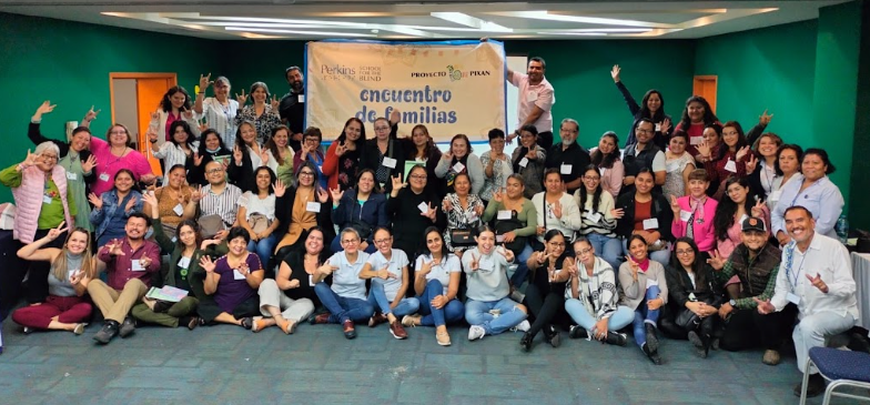 Un grupo numeroso de personas posan juntas en un interior, sonriendo y saludando a la cámara delante de una pancarta en la que se lee "Encuentro de Familias". El escenario parece ser un evento o taller comunitario.