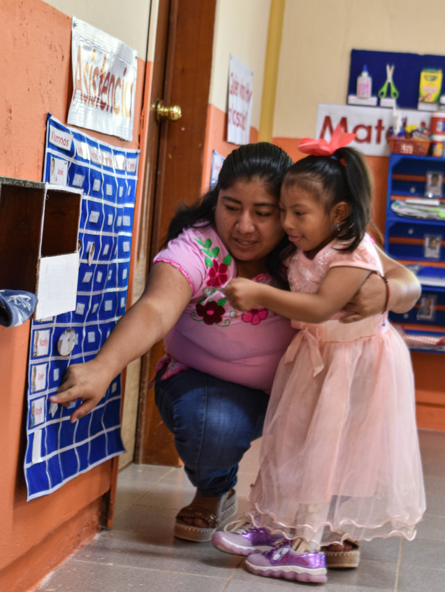 Una mujer se arrodilla junto a una niña vestida de rosa para ayudarla a colocar un elemento en una tabla de asistencia azul en un aula decorada con material educativo.