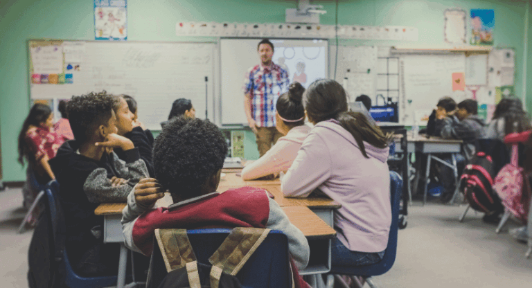 Sala de aula com vários estudantes sentados em grupos, voltados para a frente, onde um professor está em pé diante de um quadro branco e projetor. Os alunos prestam atenção ao professor, que está explicando algo. O ambiente é organizado, com cartazes nas paredes e mochilas ao lado das cadeiras