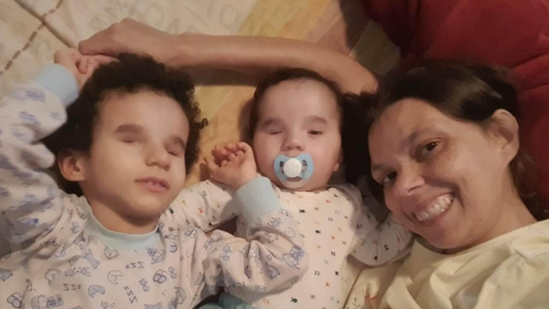 Uma mulher sorri enquanto está deitada em uma cama com duas crianças pequenas de pijama; uma criança está com uma chupeta. Elas parecem relaxadas e próximas umas das outras.