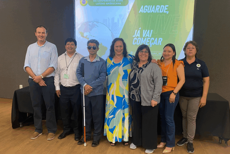 Ocho personas permanecen de pie, sonrientes, frente a una pantalla de proyector con texto en portugués. Un hombre sostiene un bastón blanco. El grupo parece estar asistiendo a una conferencia o reunión en un entorno interior formal.