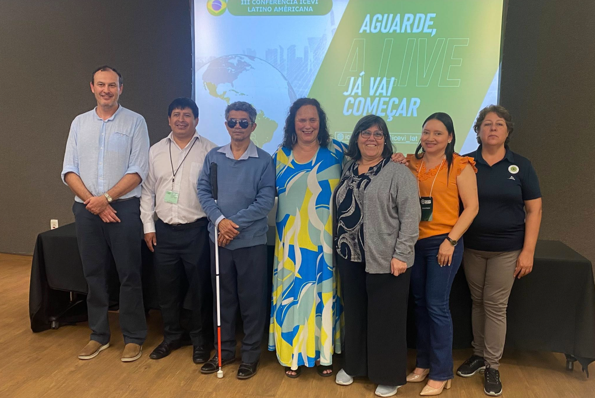 Ocho personas permanecen de pie, sonrientes, frente a una pantalla de proyector con texto en portugués. Un hombre sostiene un bastón blanco. El grupo parece estar asistiendo a una conferencia o reunión en un entorno interior formal.