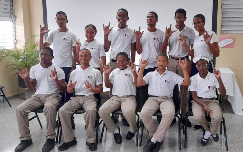 Dos filas de estudiantes adolescentes, la primera fila sentada en sillas y la segunda fila de pie, posan dentro de un aula sosteniendo el signo ‘Te quiero’ en Lengua de Señas Dominicana. Todos llevan uniformes idénticos para una escuela en la República Dominicana con camisas de cuello blanco y pantalones caqui.