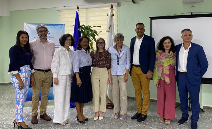 Un grupo de nueve personas permanecen de pie en el interior, sonriendo para una foto delante de unas banderas y una pantalla de proyección. Visten ropa de negocios o informal. El escenario parece ser un evento formal o profesional.