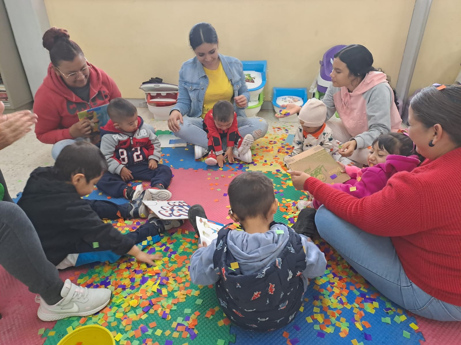 Un grupo de niños pequeños y adultos se sientan en círculo sobre colchonetas de colores, jugando con piezas de espuma multicolor y libros. El ambiente parece animado y cautivador en un interior luminoso.