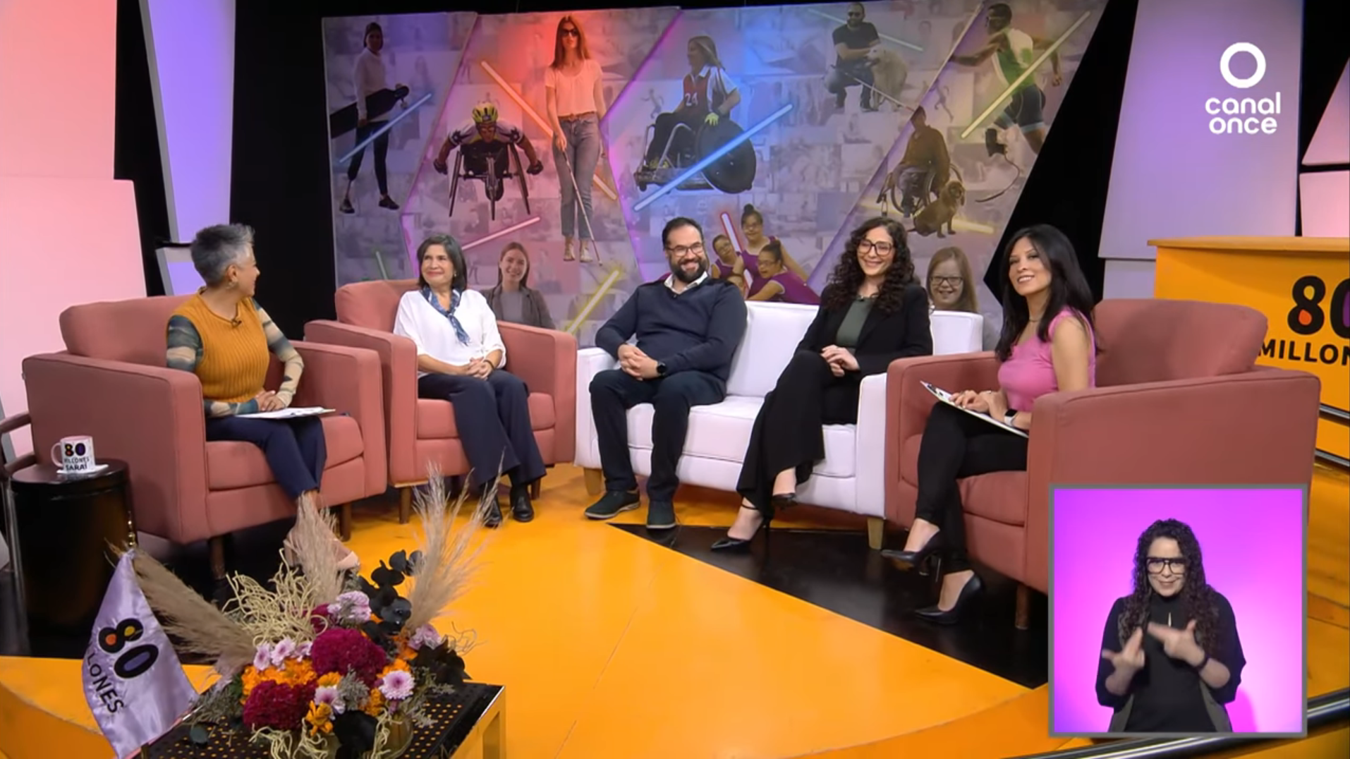 Seis personas se sientan sonrientes en un televisor brillantemente iluminado con sillas rosas y un sofá blanco. Un gran mural decora el fondo. Una mujer ofrece interpretación en lengua de signos en la esquina inferior derecha.