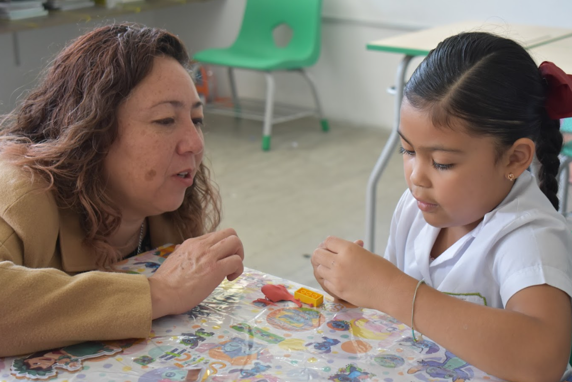 Andrea Rocha Jove se inclina para hablar con una niña que está concentrada en unos bloques de colores en una mesa. La escena parece transcurrir en un aula con una silla verde y pupitres al fondo.