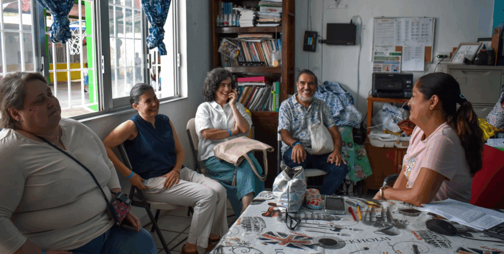 Cinco adultos se sientan juntos en un despacho, inmersos en una alegre conversación. Libros, papeles y objetos personales están a la vista por toda la habitación, creando un ambiente cálido.