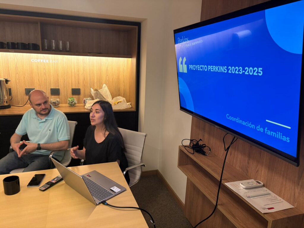Flavia da una presentación mientras un hombre, Sebas, escucha. En una pantalla se lee "Proyecto Perkins 2023-2025, Coordinación de familias". Sobre la mesa hay un ordenador portátil y documentos.