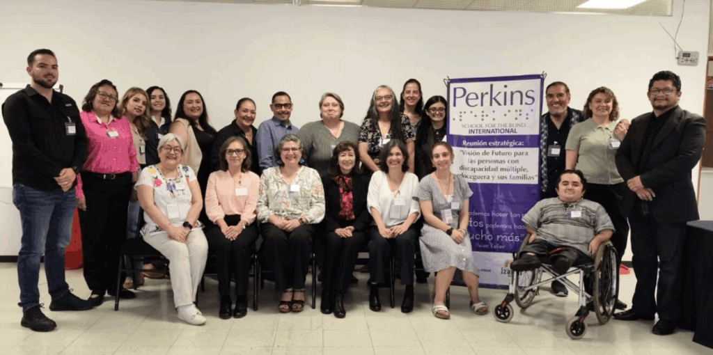 El grupo Izamal, un grupo de unas 20 personas, algunas sentadas y otras de pie, posan juntas en el interior junto a una pancarta azul de Perkins Internacional. Todos sonríen a la cámara.
