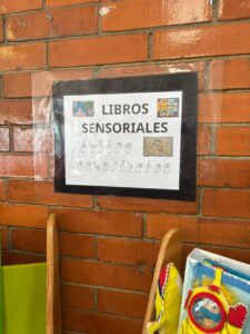 Cartel pegado en una pared de ladrillo con el título “Libros sensoriales”. Debajo del texto hay una línea con letras del alfabeto en lengua de señas.