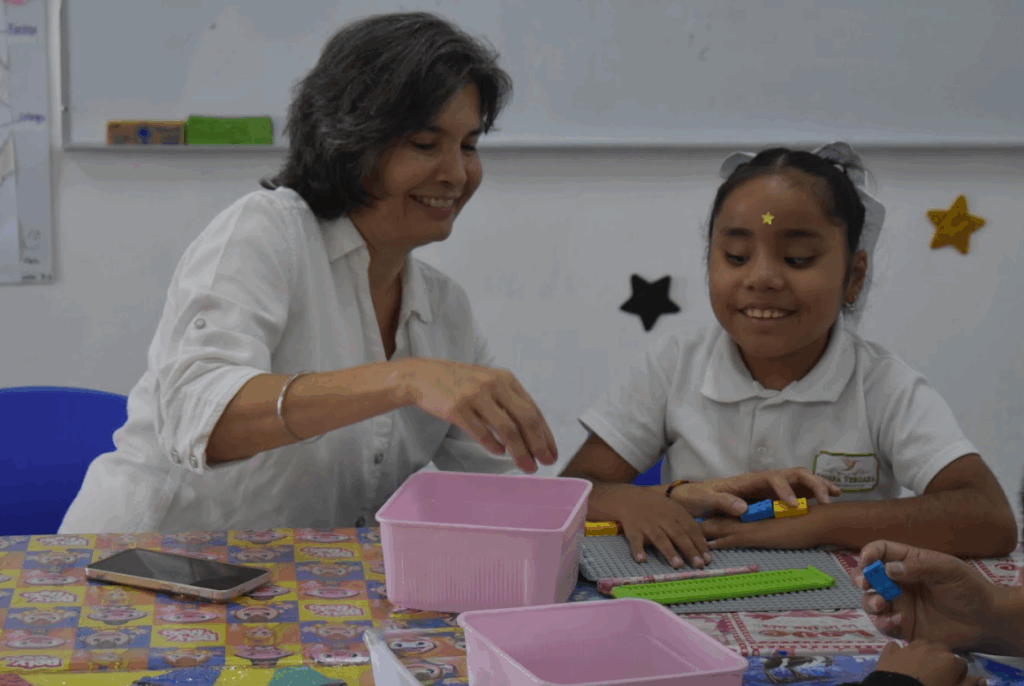 Marta Elena y una niña sentadas a una mesa con bloques de construcción y recipientes de colores, sonríen e interactúan en un aula decorada con adhesivos murales en forma de estrella.