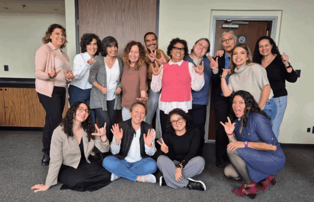 Un grupo de catorce personas, algunas de pie y otras sentadas en el suelo, sonríen y se hacen el seño de mano de 'Te quiero' mientras posan juntas en un ambiente informal.