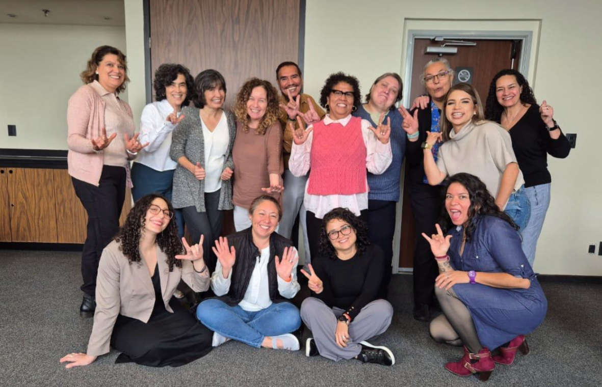 Un grupo de catorce personas, algunas de pie y otras sentadas en el suelo, sonríen y se hacen el seño de mano de 'Te quiero' mientras posan juntas en un ambiente informal.