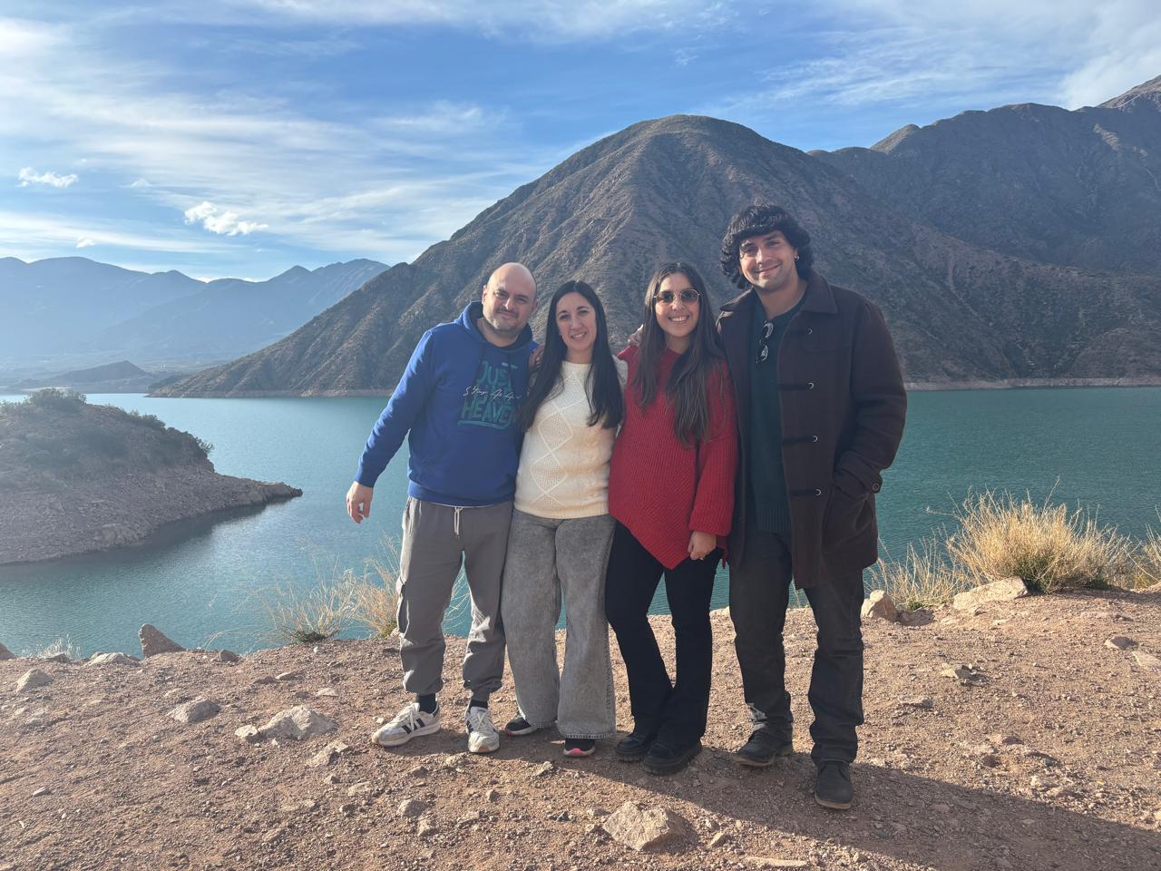 Cuatro personas están muy juntas, sonriendo frente a un lago con montañas de fondo bajo un cielo parcialmente nublado. Están vestidas con ropa de abrigo informal y parecen estar disfrutando de un lugar pintoresco al aire libre.