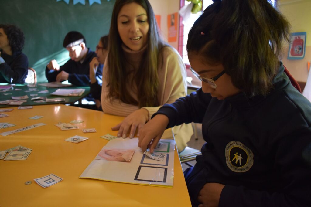 Emilce se sienta junto a un alumno que señala un dibujo en un cuaderno de ejercicios sobre una mesa amarilla. Otros alumnos trabajan al fondo en un aula con material educativo esparcido.