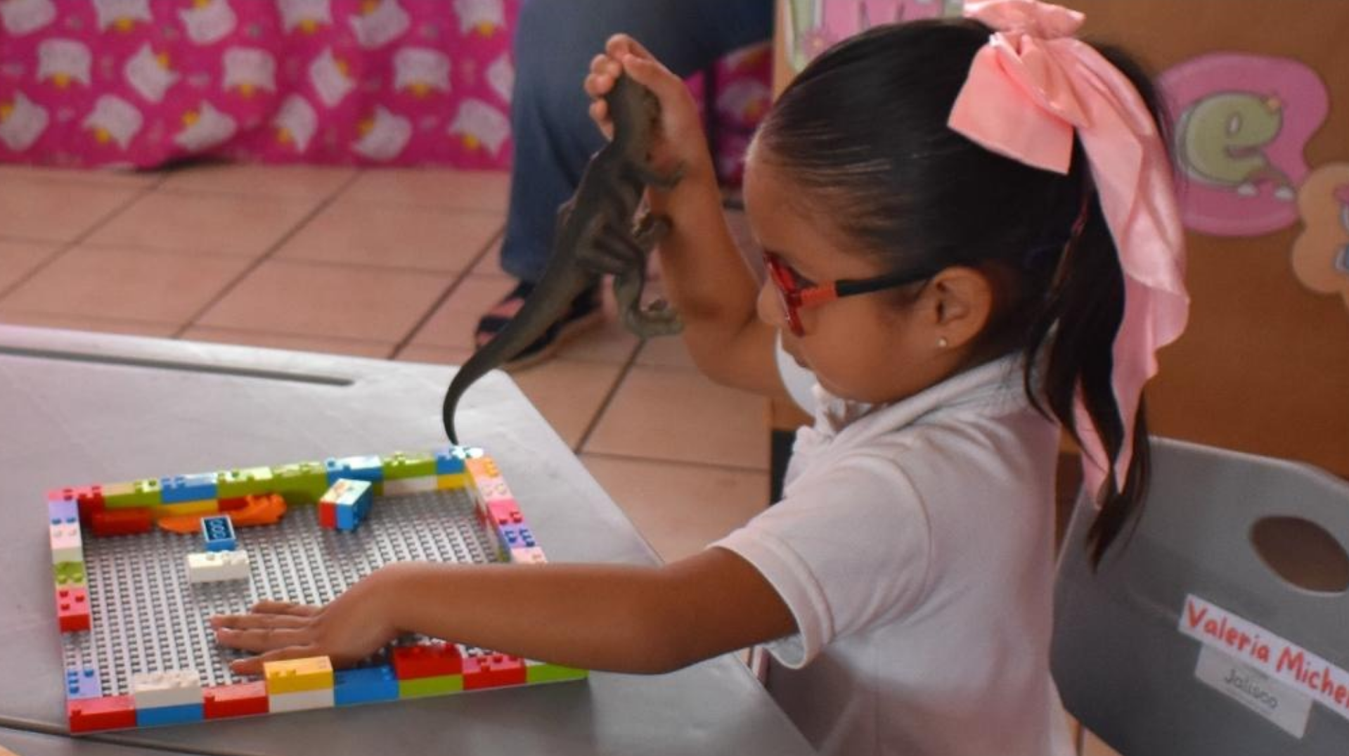 Una niña con gafas y un lazo rosa juega con un dinosaurio de juguete y bloques de colores en la mesa de una clase.