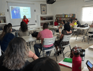Una mujer realiza una presentacion, los asistentes están sentados alrededor de mesas.