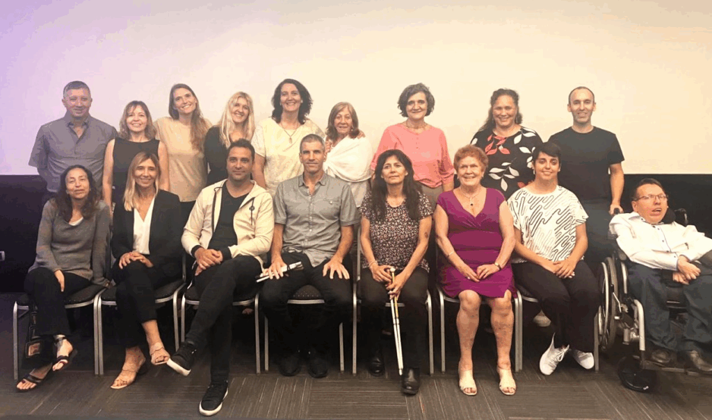 Un grupo de 17 adultos, incluidos hombres y mujeres, algunos con discapacidad, posan juntos en un espacio interior frente a una pared blanca, sonriendo a la cámara.