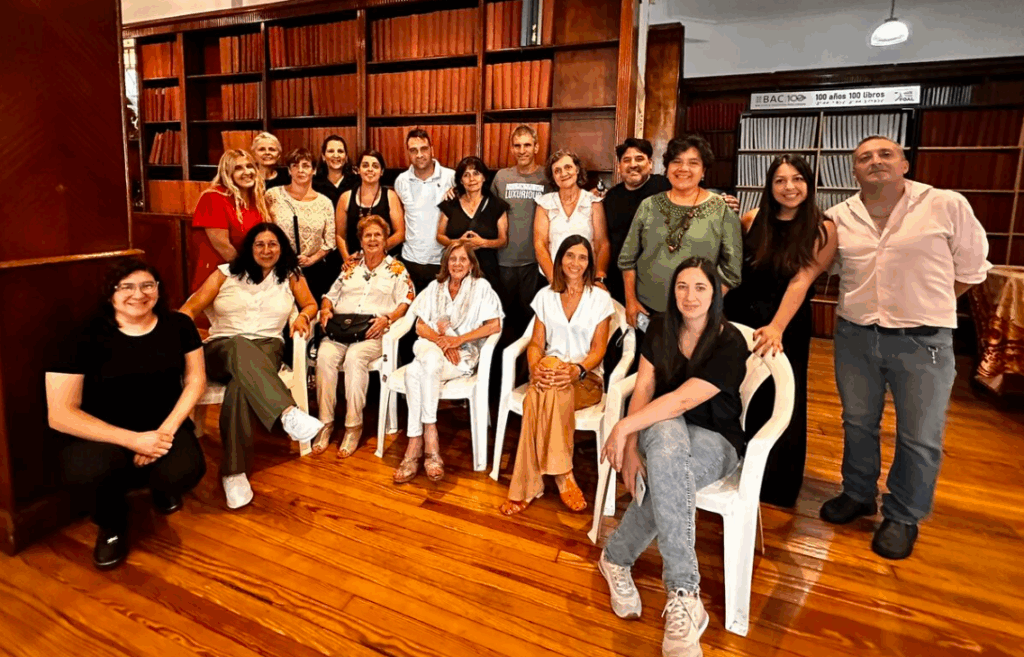 Un grupo de personas posan juntas en el interior, algunas sentadas y otras de pie, frente a estanterías repletas de libros. El escenario tiene suelos de madera y un ambiente cálido y acogedor.