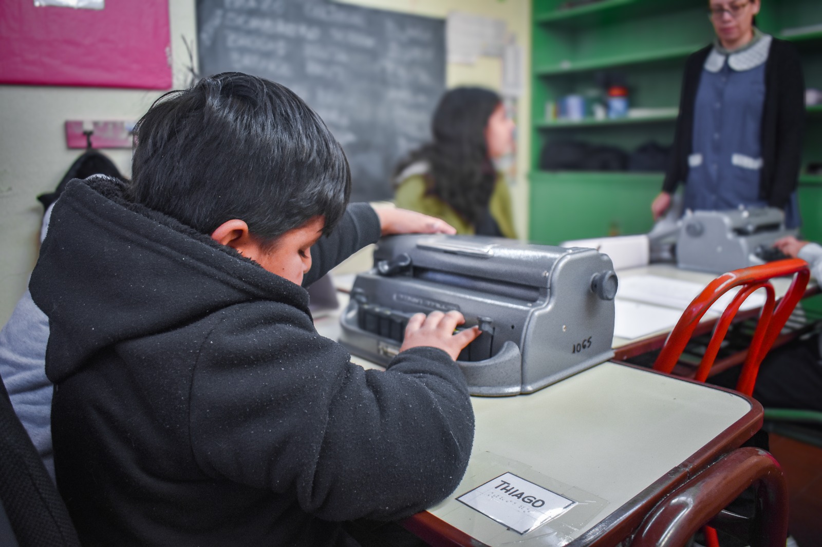 Um menino usa uma máquina de escrever em Braille em uma mesa de sala de aula. Outros alunos e um professor podem ser vistos ao fundo, perto de um quadro-negro e de prateleiras verdes.