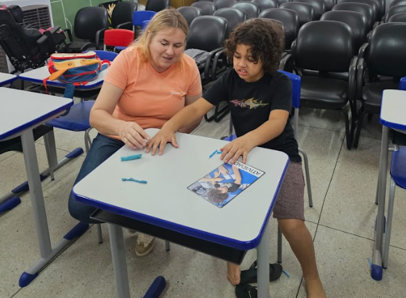 Uma professora com cabelo loiro e uma camisa laranja está sentada à mesa com uma criança, e estão a fazer uma atividade com uma fotografia e tiras de papel. O cenário é uma sala de aula, com muitas cadeiras vazias atrás delas.