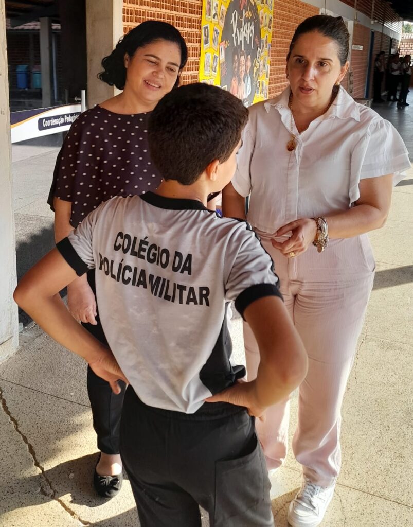 Duas professoras conversam com uma criança vestindo uniforme escolar, que está de costas para a câmera.