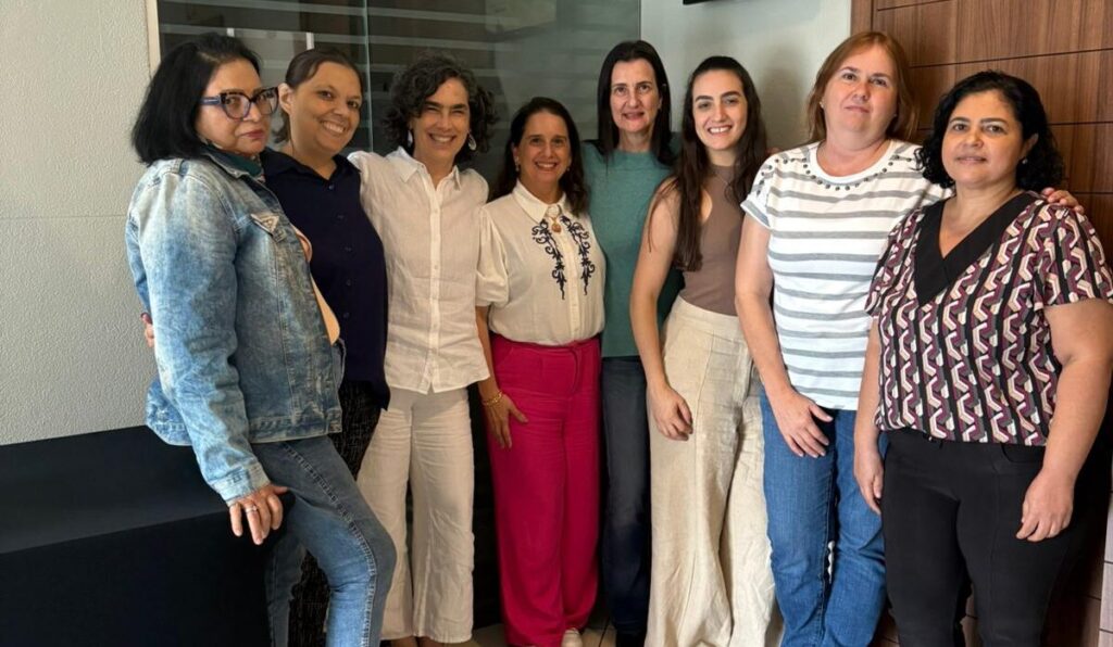 Um grupo de oito mulheres está reunido em um ambiente interno, sorrindo para a câmera. Elas estão vestidas casualmente e posam em frente a uma porta de madeira e a uma parede de vidro.
