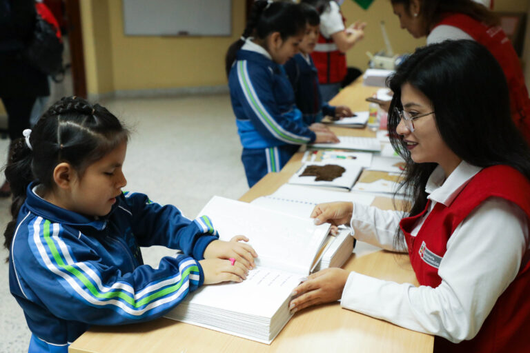 Una niña lee un libro en braille con las manos mientras una mujer con chaleco rojo la ayuda a cruzar la mesa de una clase. Otros niños y adultos también interactúan en el fondo.