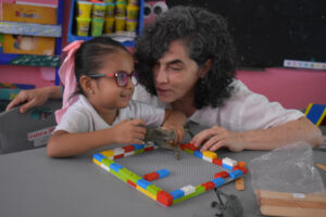 Una niña y una mujer adulta juegan juntas en una mesa, construyendo con ladrillos LEGO de colores y sujetando animales de juguete en un aula. La mujer sonríe cariñosamente mientras interactúan.