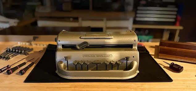 En un taller bien iluminado, sobre una mesa de madera rodeada de herramientas y un maletín de madera, se encuentra una Perkins Brailler.