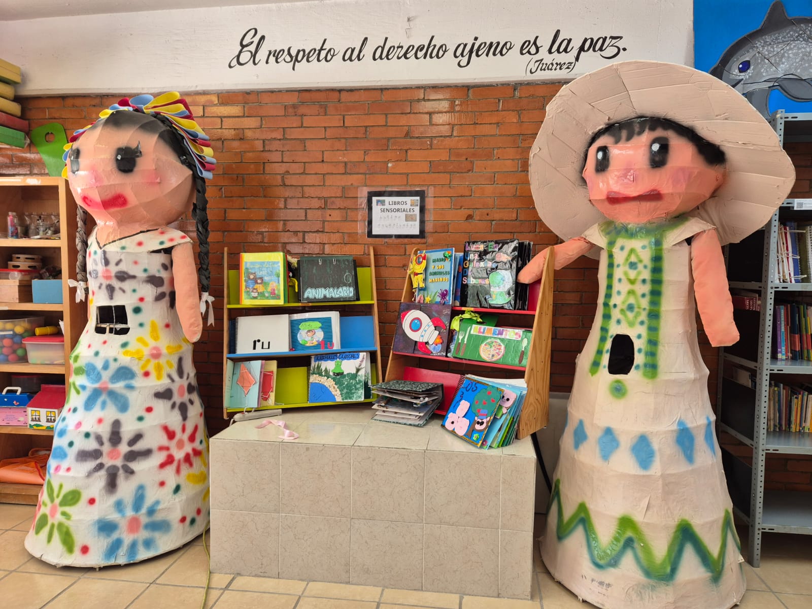 Dos grandes muñecas de papel maché están junto a una estantería llena de coloridos libros infantiles.