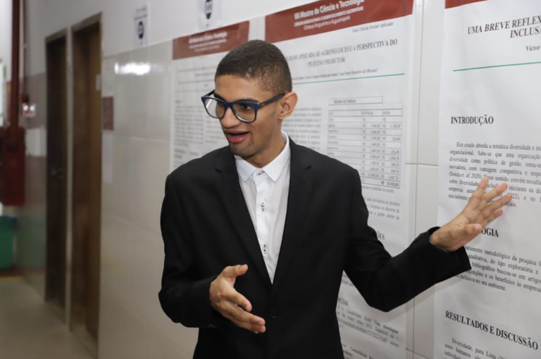 Un joven con traje y gafas gesticula mientras hace una presentación frente a una pared con carteles y textos académicos. Parece estar explicando los resultados de una investigación o un proyecto en un entorno educativo.