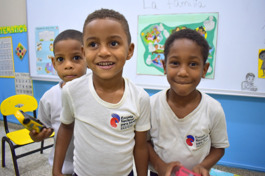 Tres chicos jóvenes con uniformes escolares blancos sonríen y posan juntos en un aula, frente a una pizarra blanca con dibujos de colores y carteles educativos en las paredes.