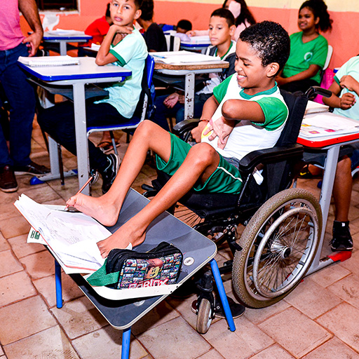 Um menino em cadeira de rodas escreve em um caderno usando o pé em sua mesa em uma sala de aula, cercado por outros alunos que também estão trabalhando.