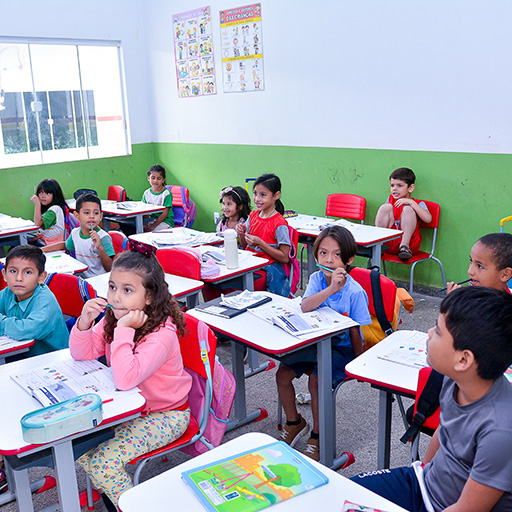 Um grupo de crianças pequenas está sentado em carteiras em uma sala de aula bem iluminada, ouvindo atentamente. A sala de aula tem paredes verdes e brancas, pôsteres educativos e grandes janelas que permitem a entrada de luz natural.